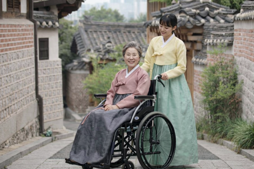Elderly mother in pink hanbok seated in a folding wheelchair with her adult daughter walking beside her in hanbok, both smiling - — wheelchair rental for elderly parents in Seoul.